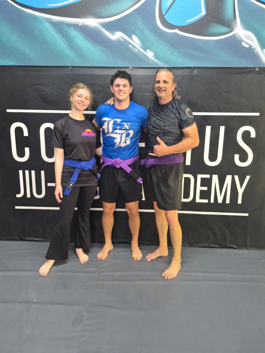 Three teammates wearing blue and purple belts in front of the academy banner