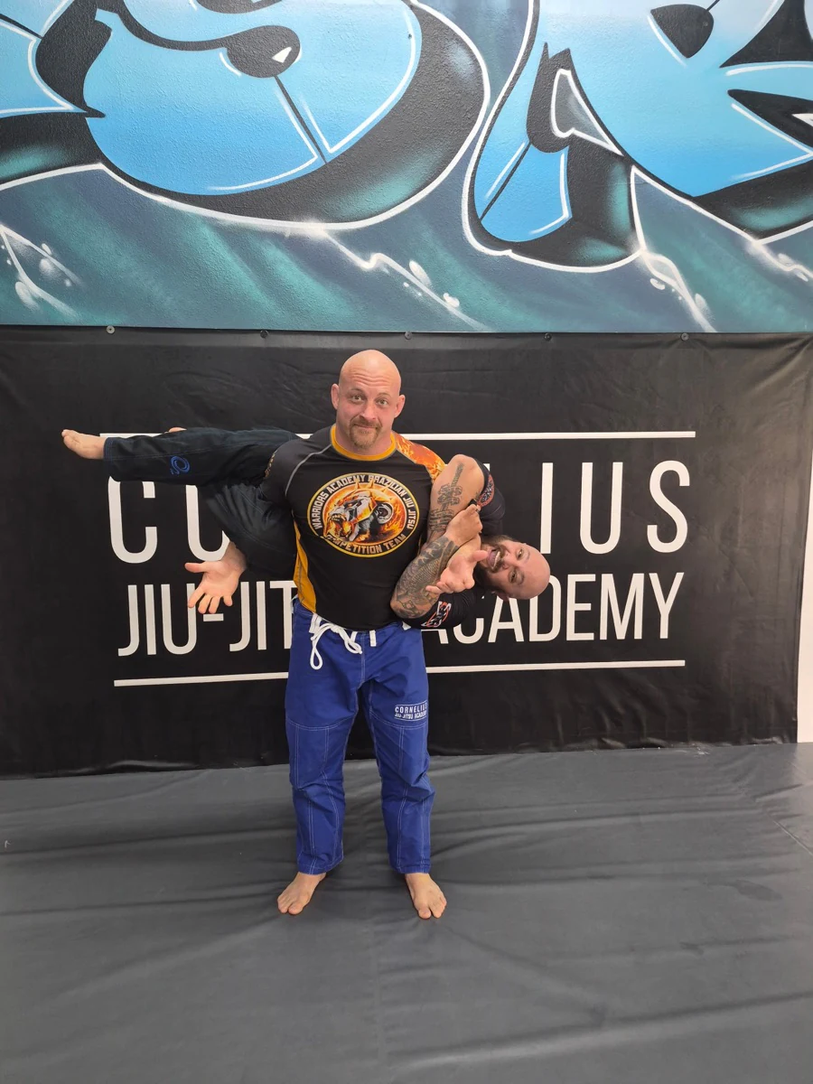 Two teammates playfully posing with one carrying the other in front of the academy banner