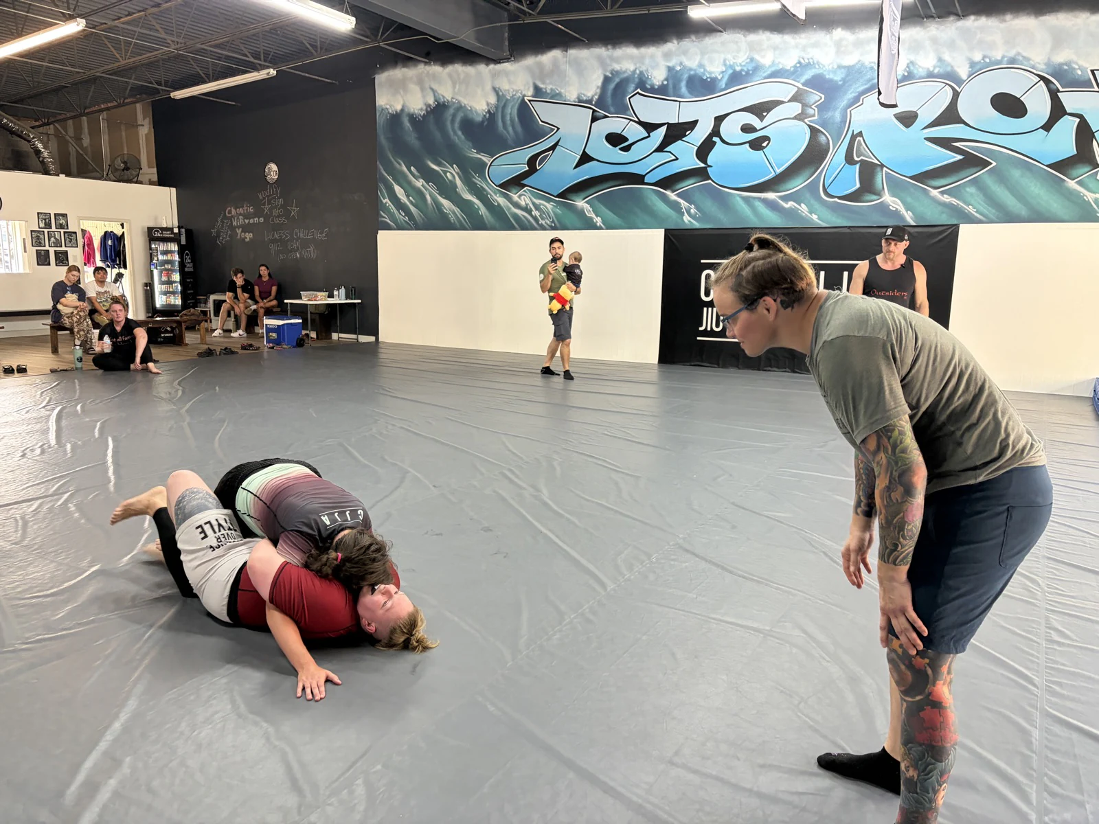 Two training partners grappling while another person watches beside the mats